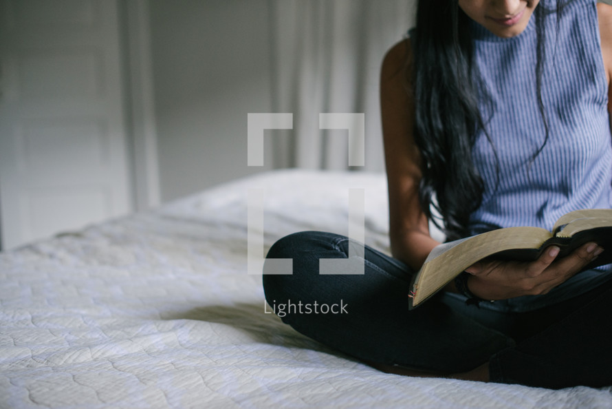 A teen girl reading the Bible while sitting on her bed.