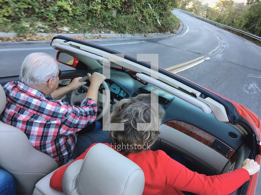 An elderly couple driving a red convertible on a curvy road 