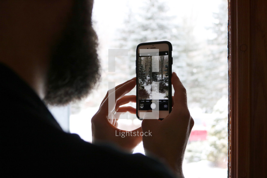 a man taking a picture of fresh snow with a cellphone while looking out a window 