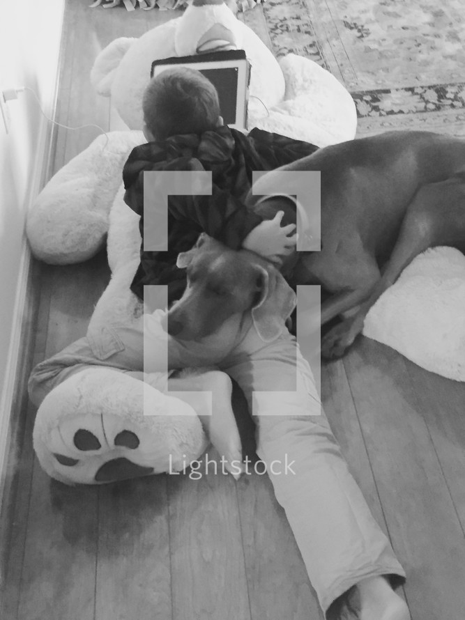A boy playing on an iPad and lying on a large teddy bear on the floor with his pet dog 