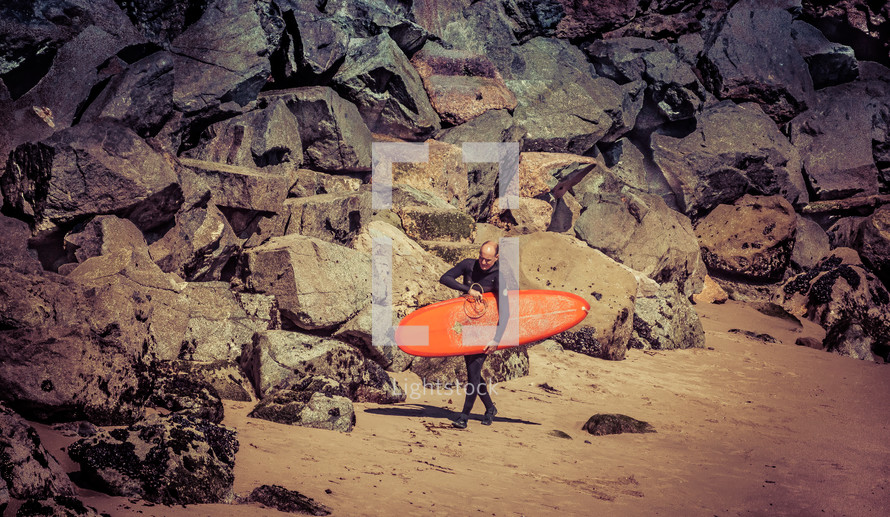 A man in a wetsuit carrying a surfboard along a beach lined with boulders.
