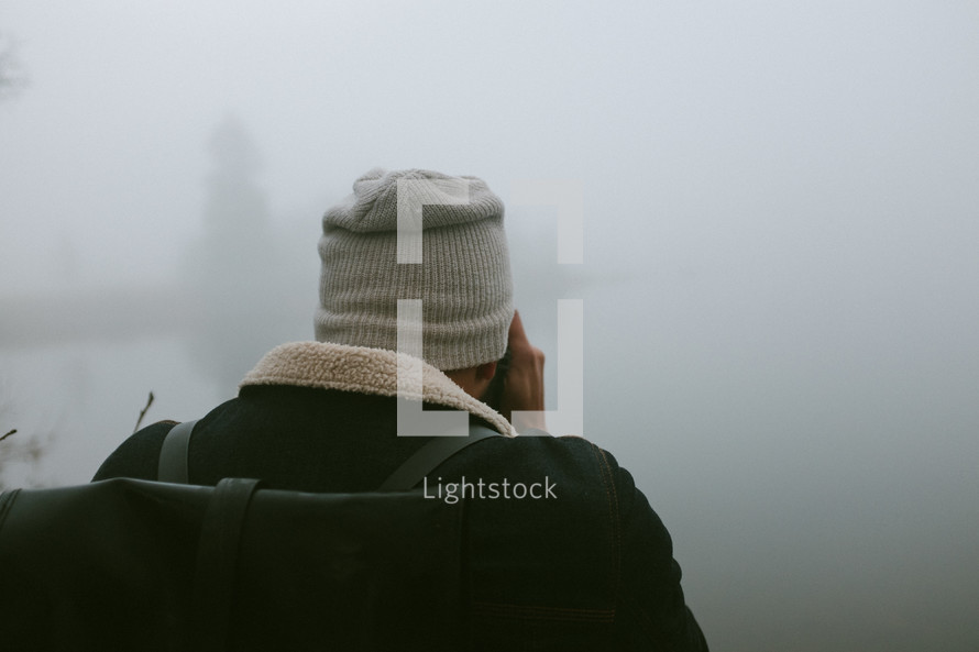 a man with a backpack standing around a foggy lake 