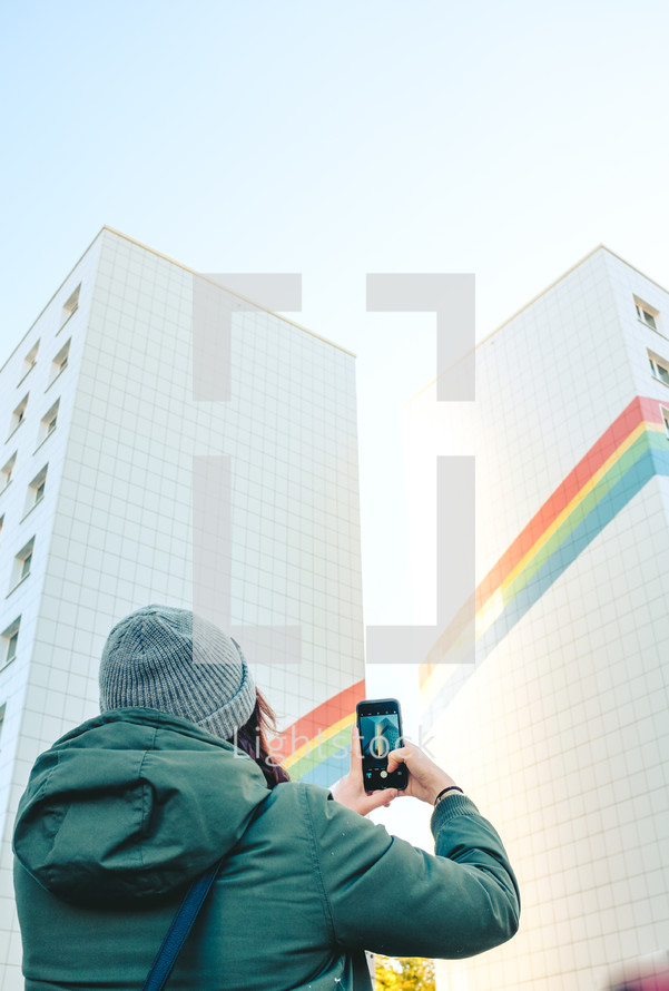 woman taking a picture of a rainbow painted on the side of a building 