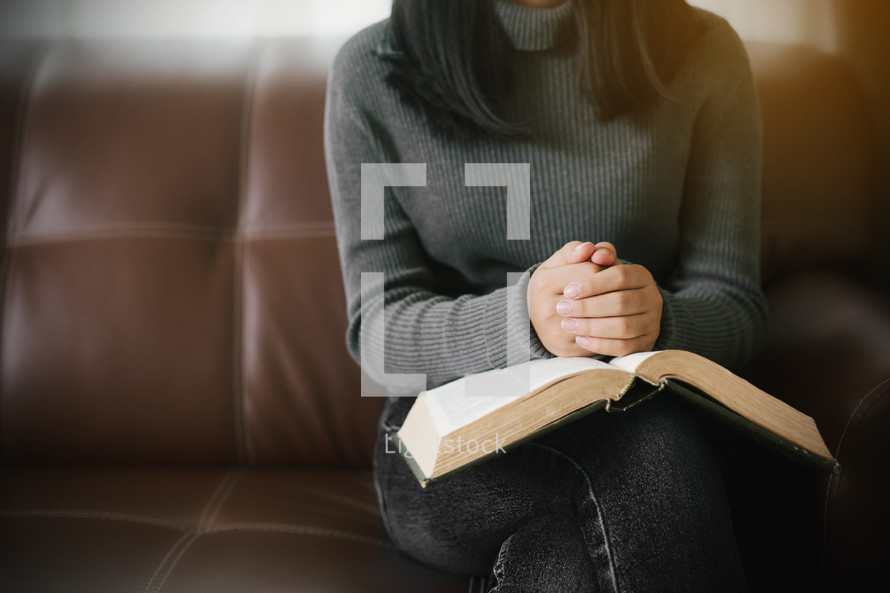 a woman sitting on a couch praying over a Bible in her lap 