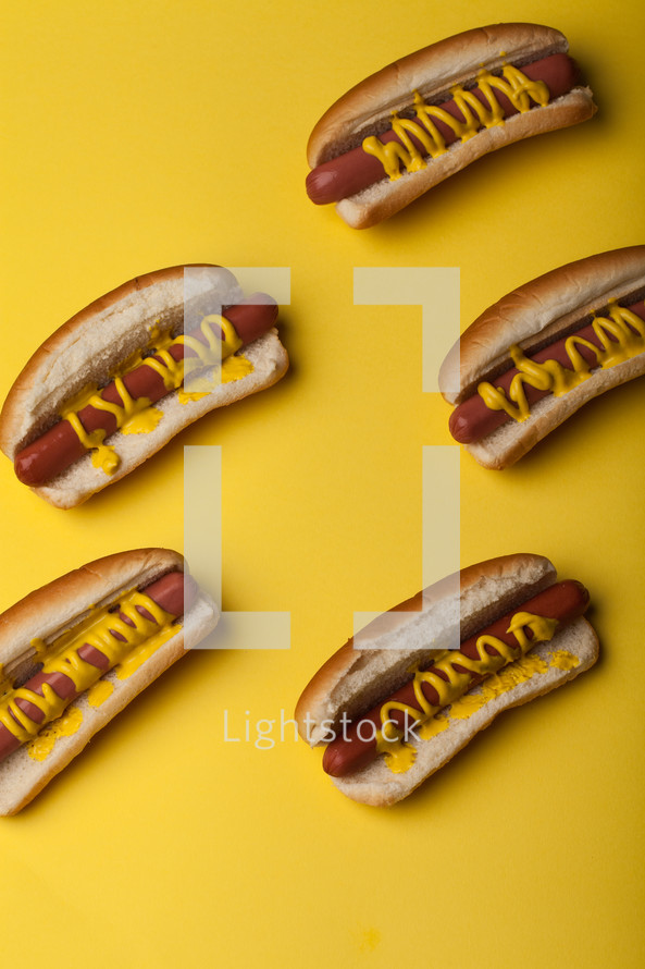 Hot dogs and buns drizzled with mustard on a bright yellow background.