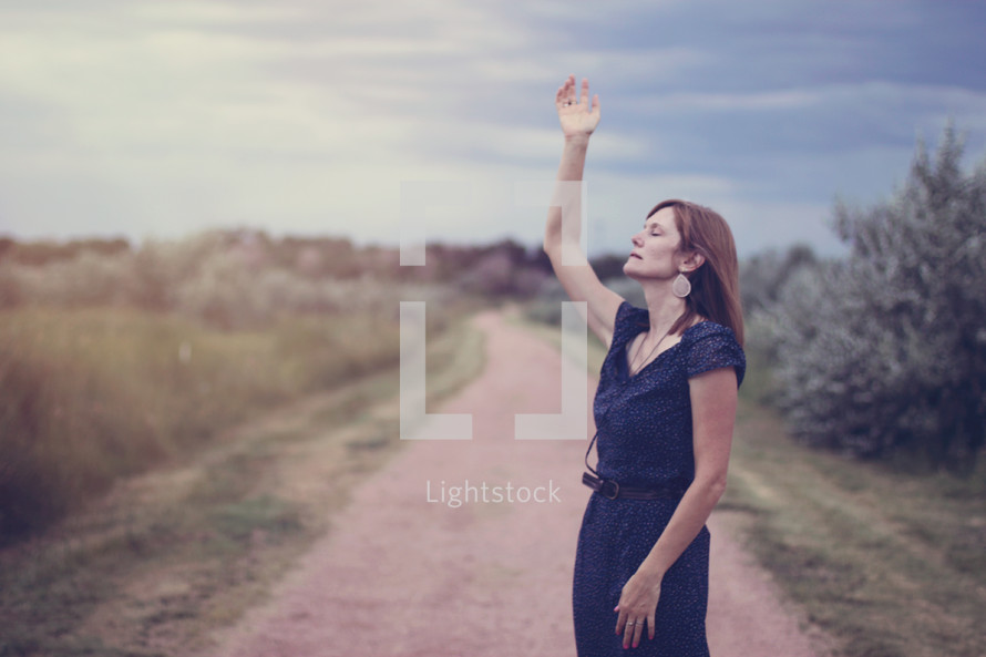 woman standing on dirt road with hand raised praising God 