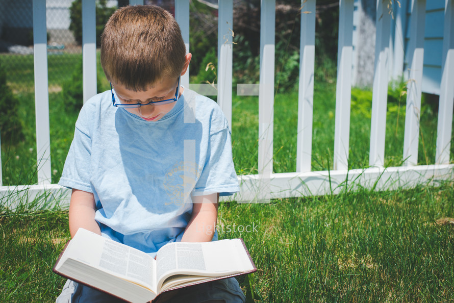 a young boy sitting in grass on a sunny day reading a Bible in solitude with his head bowed 