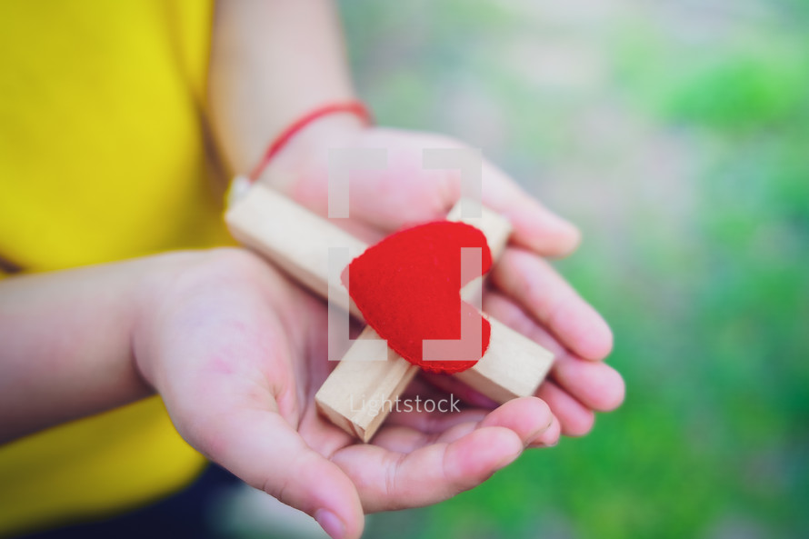 Close up hands of child girl holding cross with red heart in hand, love of jesus concept.