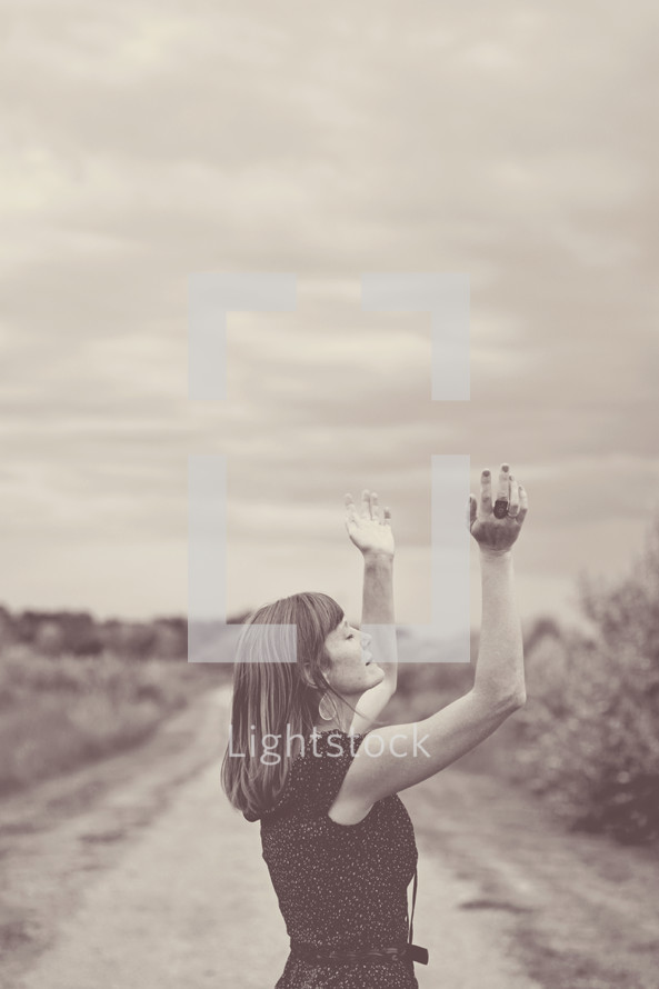 woman standing on a dirt road with hands raised praising God 