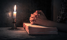 praying hands over and open Bible by candlelight 