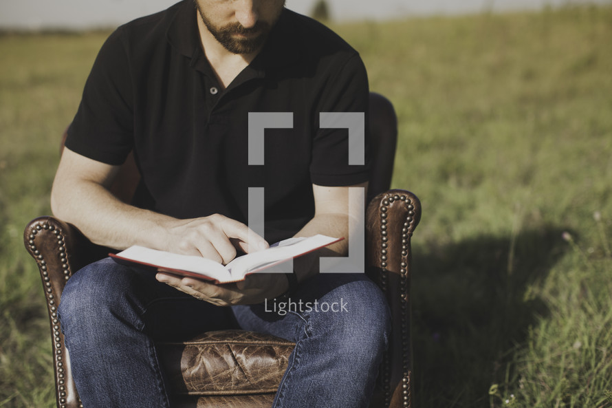 a man reading a Bible while sitting in a leather chair in a field 