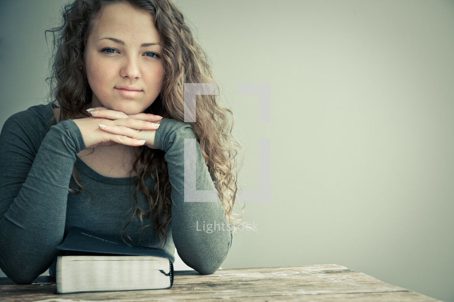 Girl with chin on folded hands with Bible on the table.