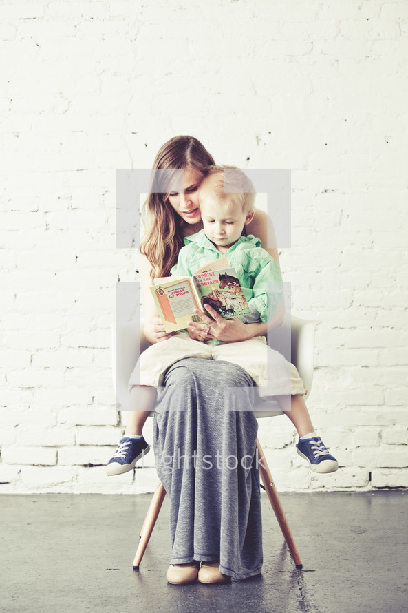 Mother holding son in her lap while sitting in a chair against a brick wall reading.