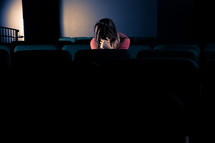 woman sitting in a dark empty church with her head bowed and her hands covering her face
