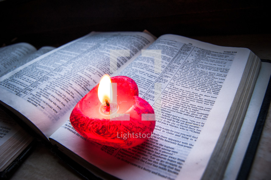 A pink heart-shaped lit candle sits upon an open bible