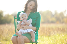 Mother holding toddler daughter while sitting in a chair in a field.
