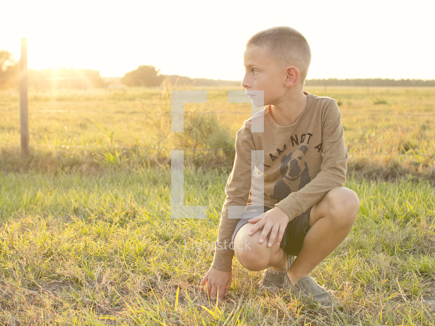 a boy squatting in a field at sunset 