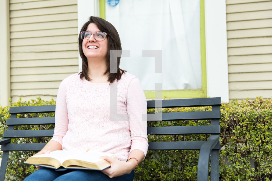 A woman looks up, smiling, while sitting on a bench with an open Bible in her lap.