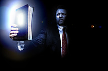 Man in suit holding shining light of the Bible up in the midst of darkness.