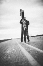 man walking in the center of a highway carrying a large cross - re-enacting Christ's walk, bearing the cross