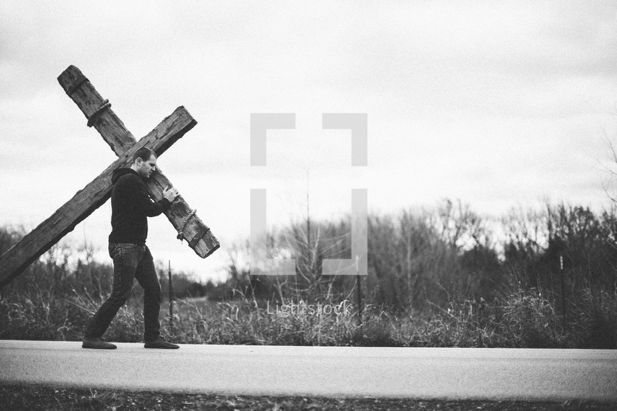 a man walking on a highway carrying a cross  - re-enacting Christ's walk, bearing the cross