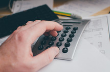 A man's hand punches in numbers on a calculator sitting atop of bills on a desk