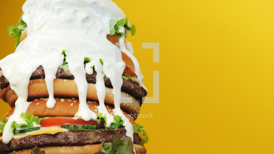 Huge fresh stacked burgers. Mayonnaise fat sauce poured from top. Yellow background. Fastfood, unhealthy food concept. Copy space.