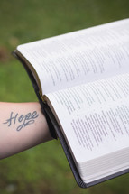 Tattooed arm holding an open Bible outside.