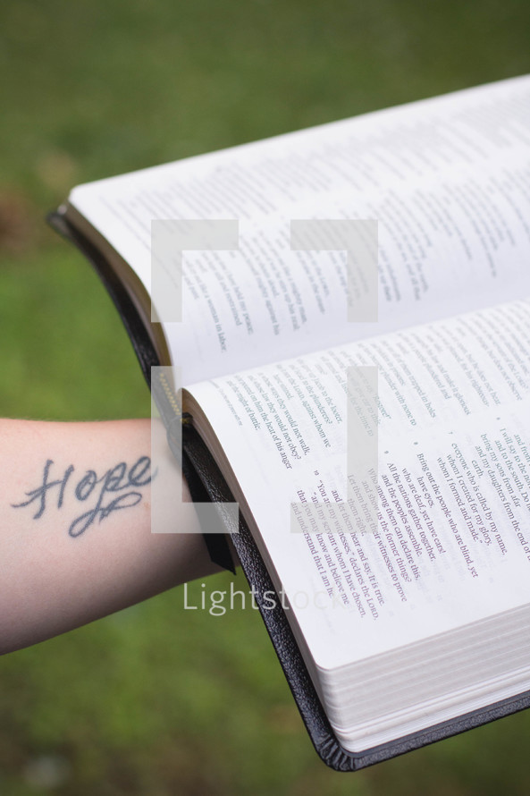 Tattooed arm holding an open Bible outside.
