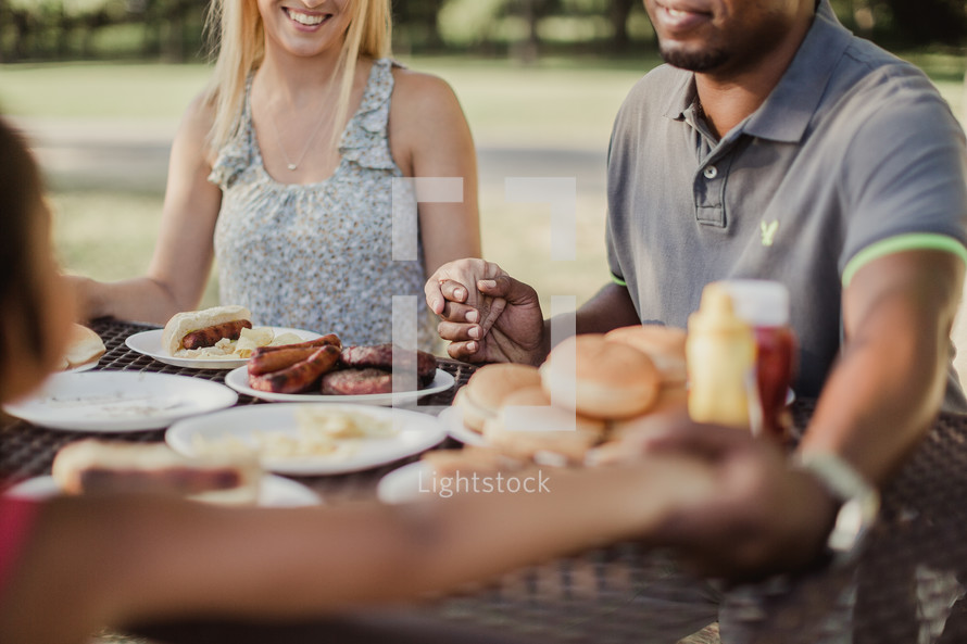 family holding hands in prayer at a picnic table outdoors 