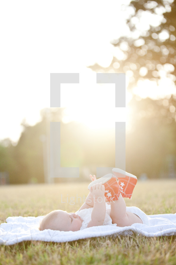 Infant laying on a blanket in the grass on a sunny day, grabbing her shoes.