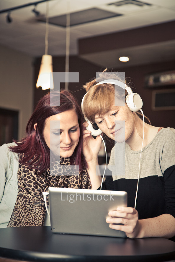 two women listening to music through headphones from an iPad
