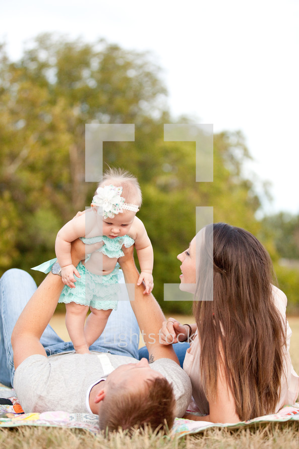 Couple laying on a blanket in the grass, playing with infant daughter.