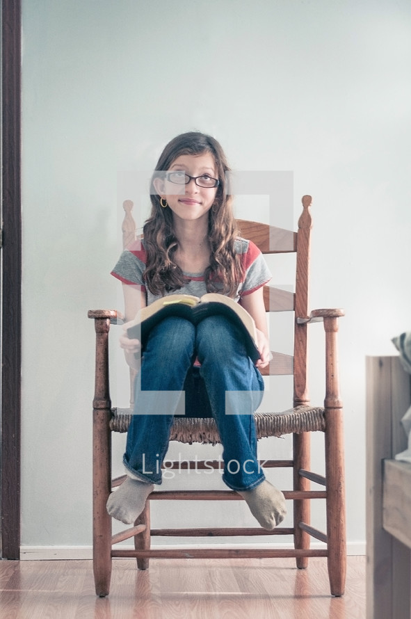 A girl sitting in a bedroom chair with an open bible, thinking.