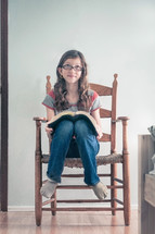 A girl sitting in a bedroom chair with an open bible, thinking.