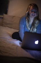 young woman sitting on her bed looking at a computer screen in a dark dorm room 