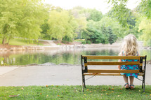 Girl sitting on a park bench by a lake surrounded by trees.