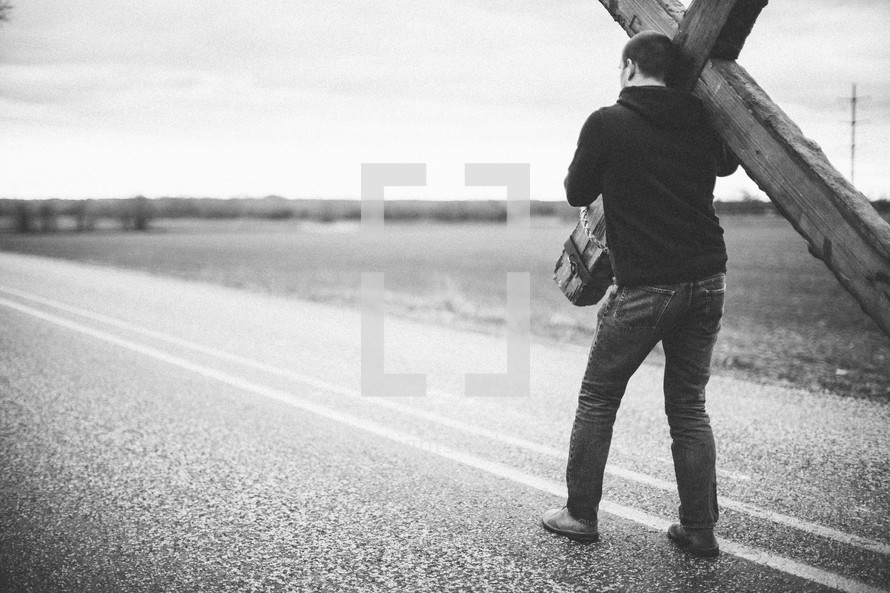 man carrying a large cross along the center lines of a highway - re-enacting Christ's walk, bearing the cross