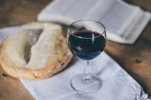 A glass of wine sits on a table next to a loaf of bread and an open bible