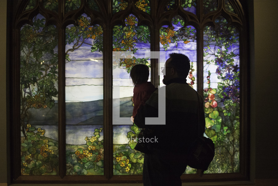 a father holding his toddler looking at a stained glass window 