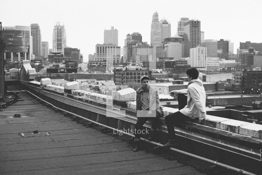 two teens sitting on the ledge of a city rooftop