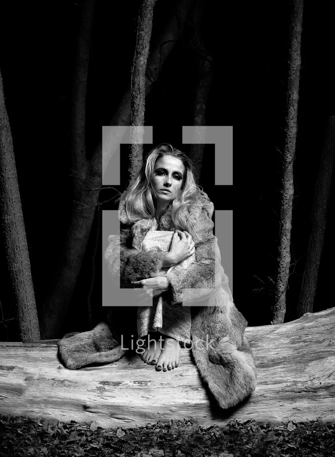 woman sitting on fallen tree wearing a fur coat and holding her legs