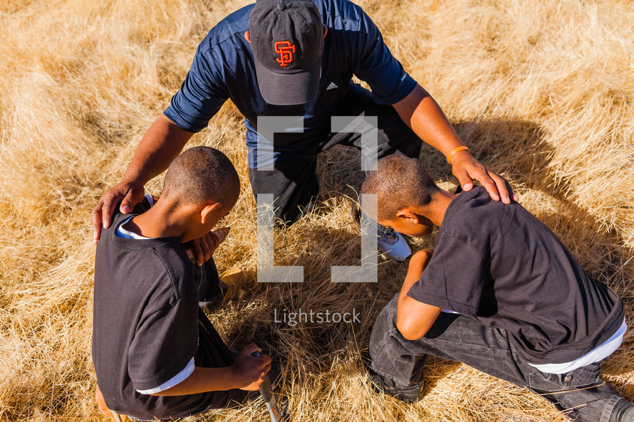 father and sons kneeling in prayer in a field african american laying hands