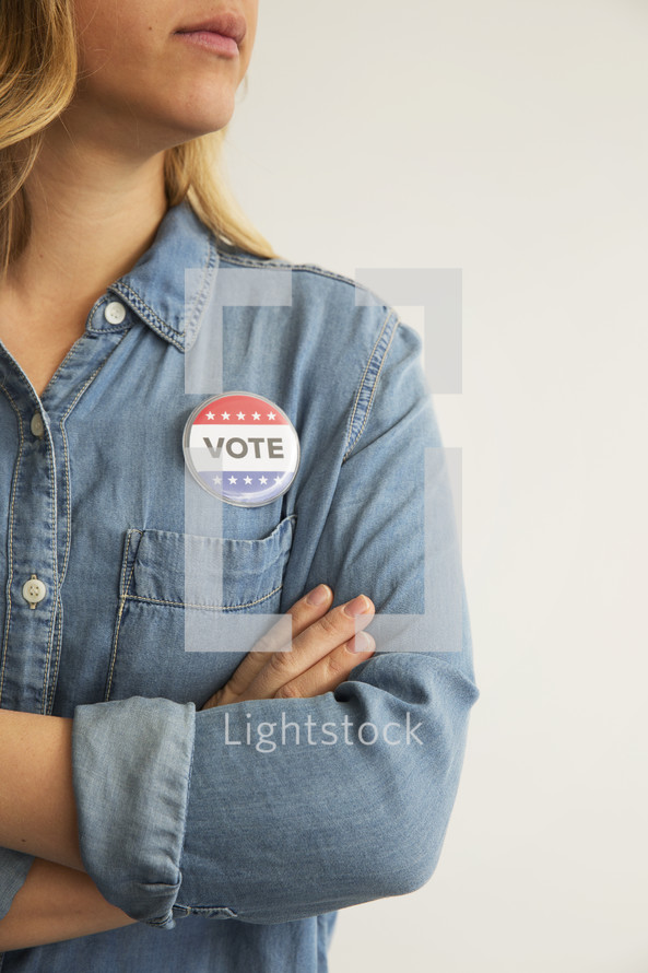 A woman with arms crossed wearing a denim shirt and a ,"Vote," button.