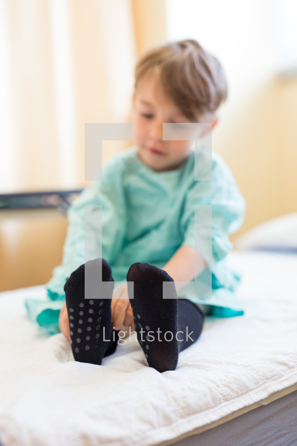 a boy child in a hospital gown and socks sitting  in a hospital bed 