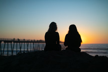 two women sitting on a beach at sunset 