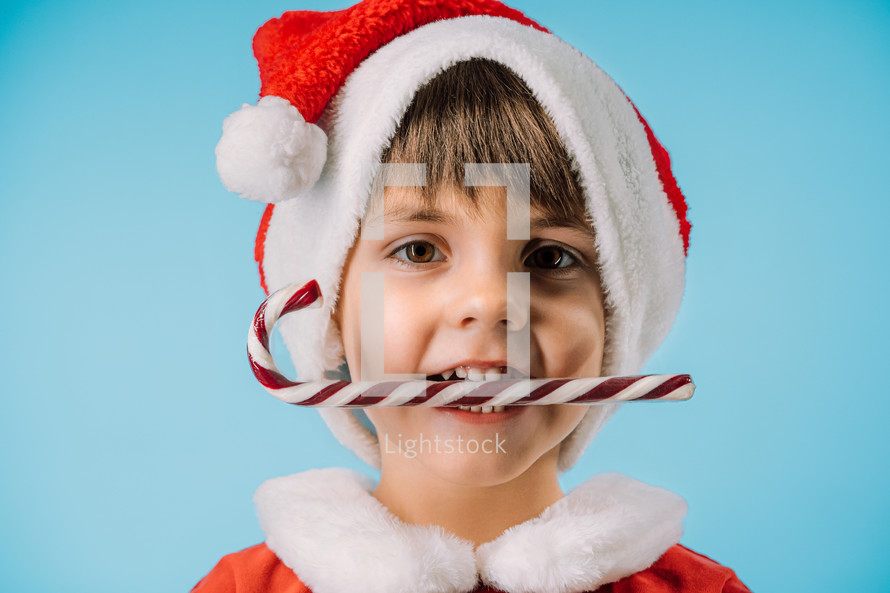 Funny boy in Santa Claus hat and sweet caramel cane on blue studio background. Christmas celebration. Happy childhood, kid, lovely son. High quality photo