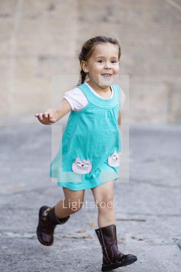 A little girl running in  a blue dress and boots.