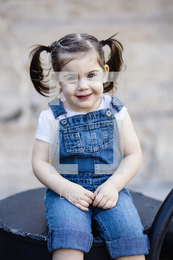 A little girl in overalls sitting down.