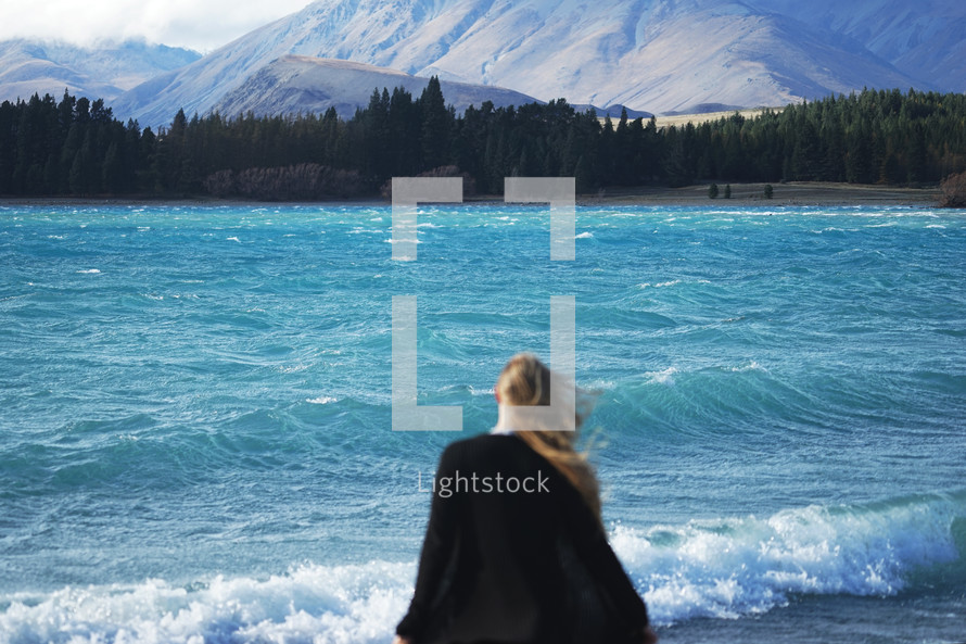 a woman standing on a shore looking out at the water and mountains 